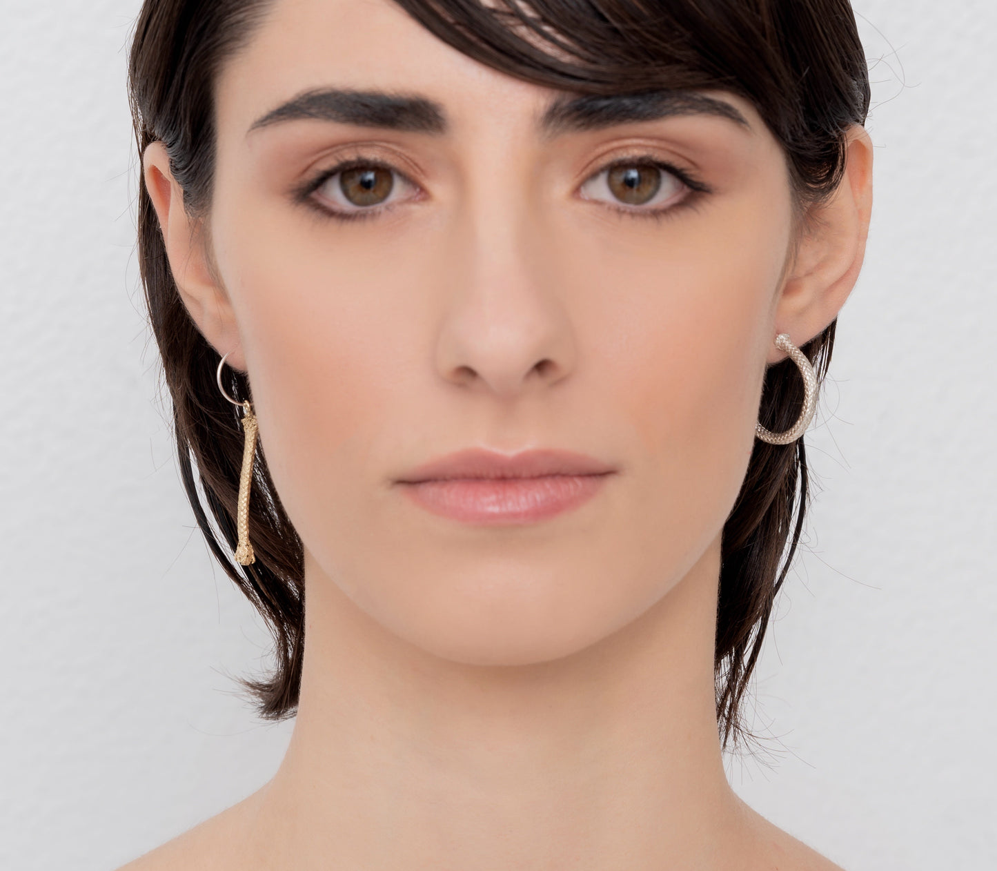 A person with short, dark hair is facing forward. They are wearing Nautical Hoop Corto Maltese Inspired Creole Earrings, made of sterling silver and featuring a rope design in size M, and they have a neutral expression. The background is plain and light-colored.