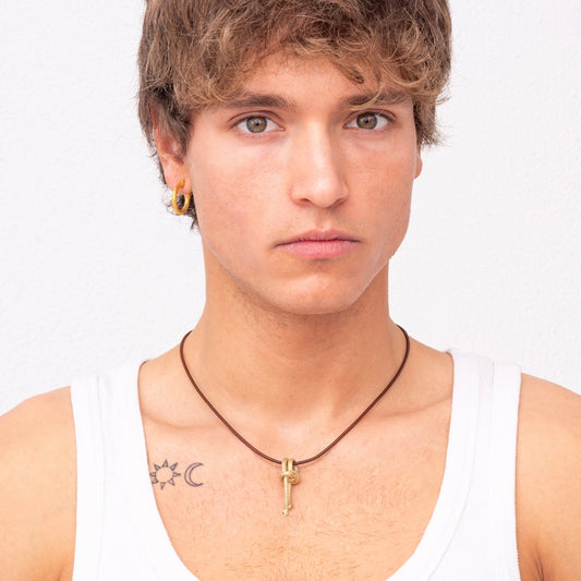 A person with short, light brown hair and blue eyes wears a white tank top, the Nautical Golden Knot Pendant - Ballestrinque S in green gold plated brass around their neck, and two earrings. Tattoos of a crescent moon and a sun are visible on their shoulders.