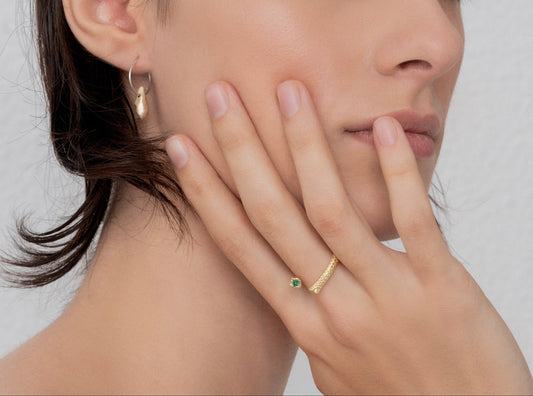 A person with short dark hair wears a 24k gold plated Golden Drop Hoop Earring Nautical Inspired and a gold ring with a green gemstone, both handmade by Angela Lago, with their hand touching their face.