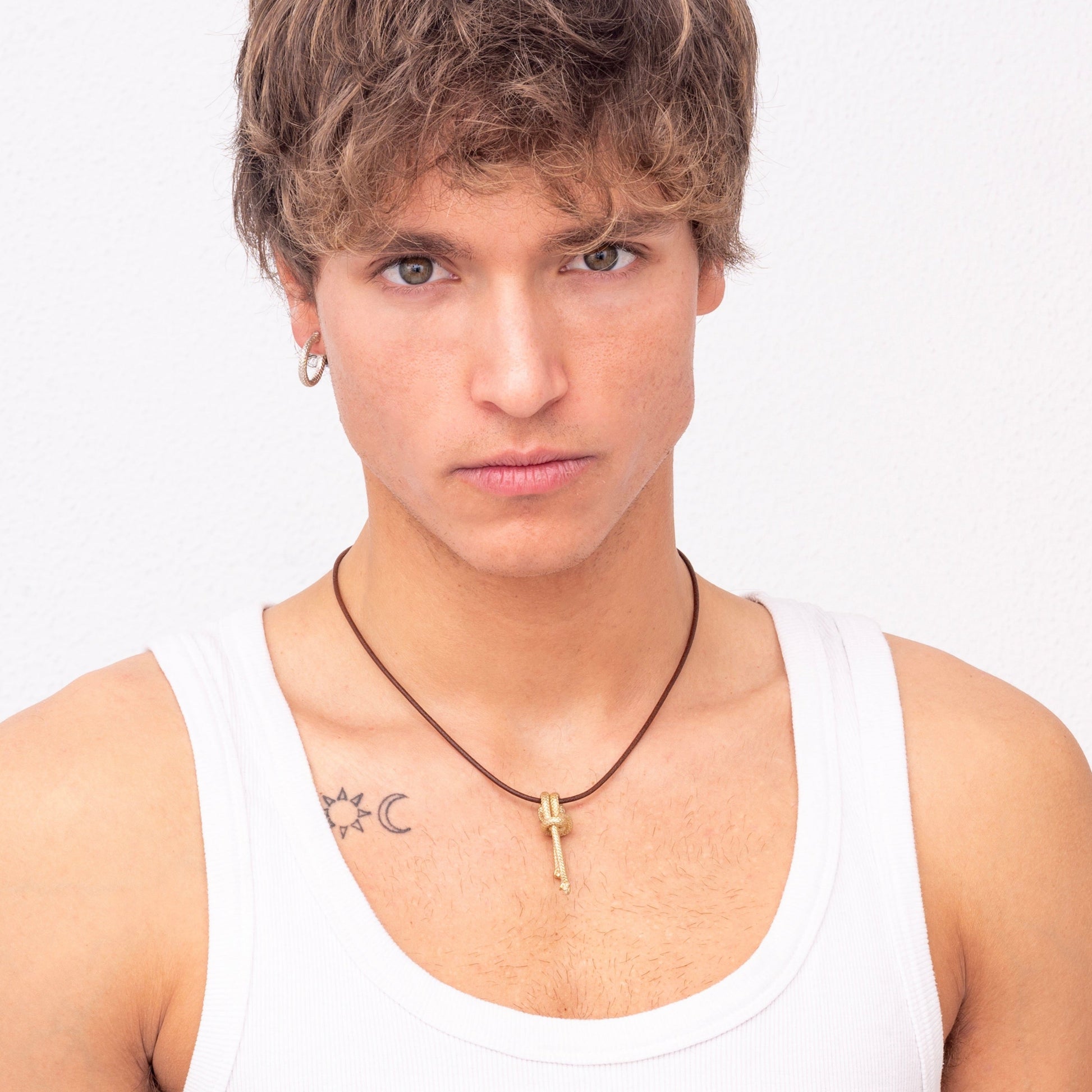 Young man with blonde hair wearing a white tank top, small hoop earrings, and a Nautical golden knot pendant - Ballestrinque S necklace, posing against a plain white backdrop. Has tattoos on both shoulders.