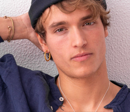 Person with wavy hair and a blue cap, wearing a blue shirt, and sporting the Nautical Hoop Corto Maltese Inspired: Creole Earring Rope Size M, looks at the camera with a neutral expression. One hand is resting behind their head.