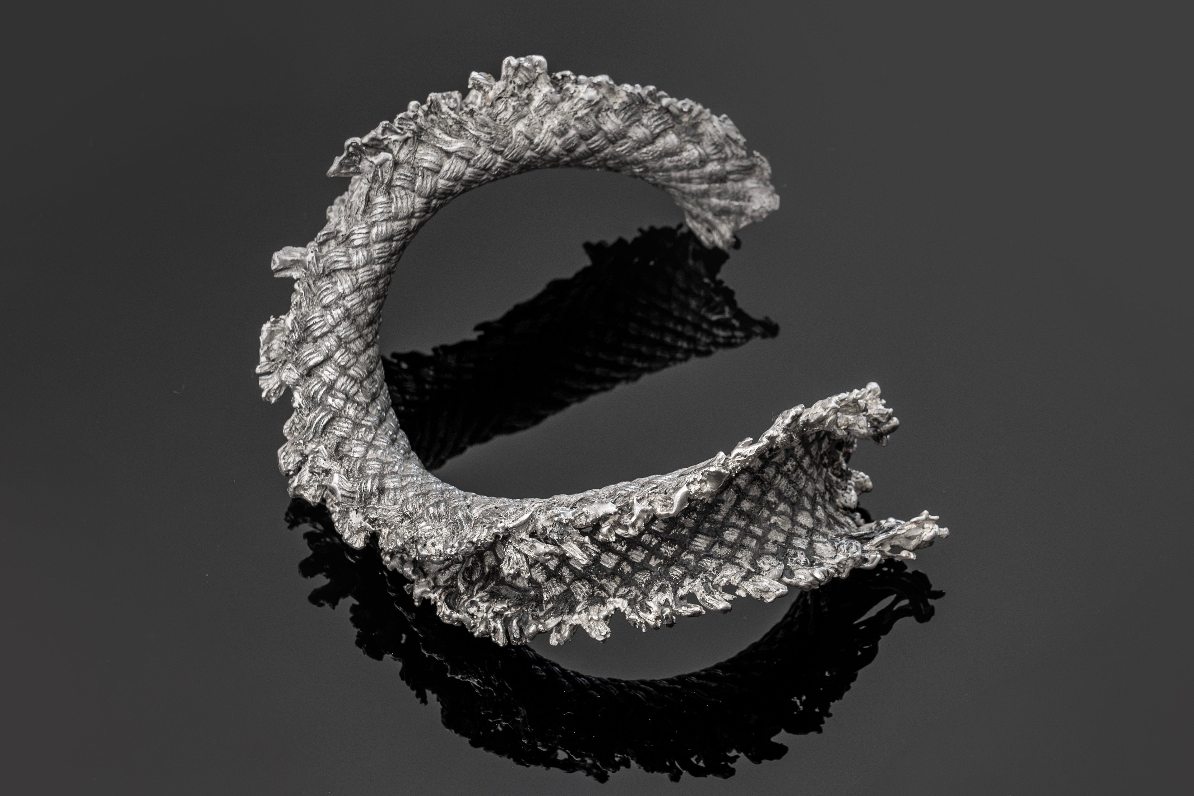 A sculptural, open-ended ring-shaped object with a textured, scale-like surface in silver-gray, shown on a dark gray reflective background with a black reflection below.