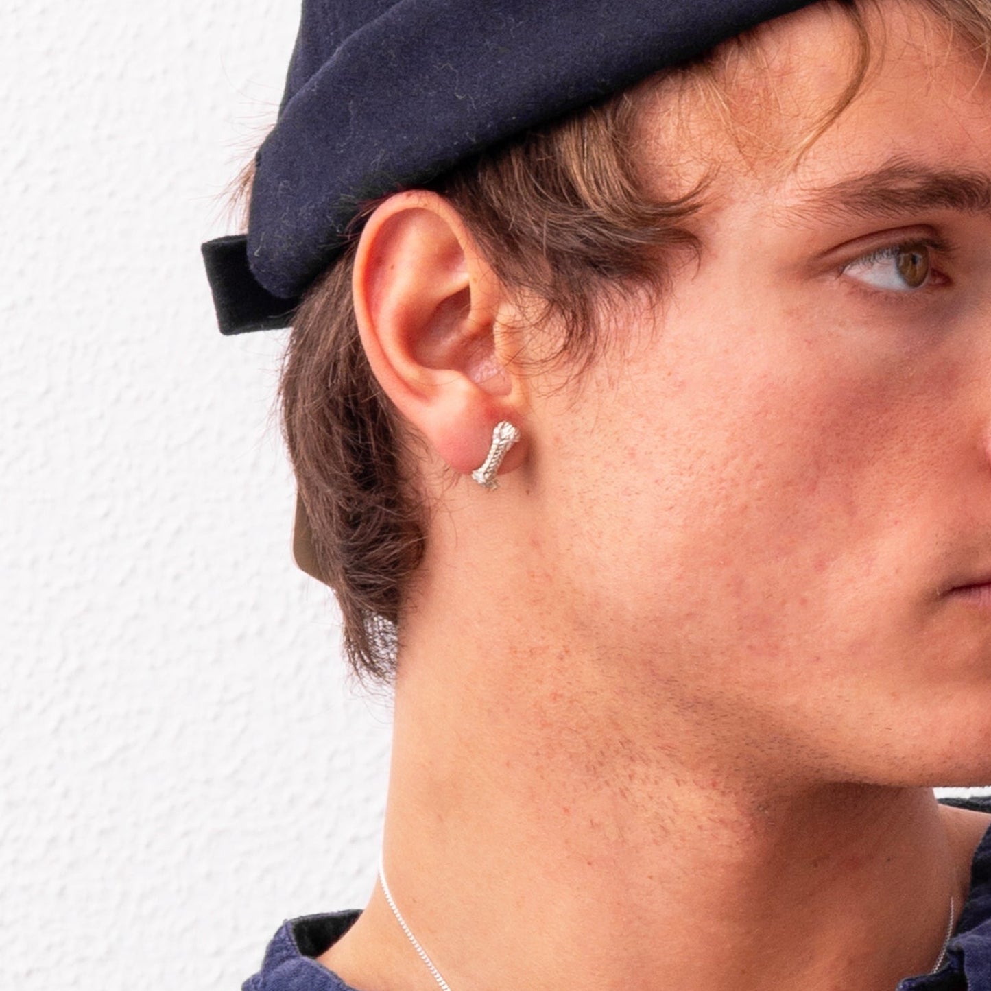 A person with light brown hair wearing a dark rolled-up beanie and a Sterling Silver Rope S Earring: Handmade Nautical Jewel looks to the side against a white background.