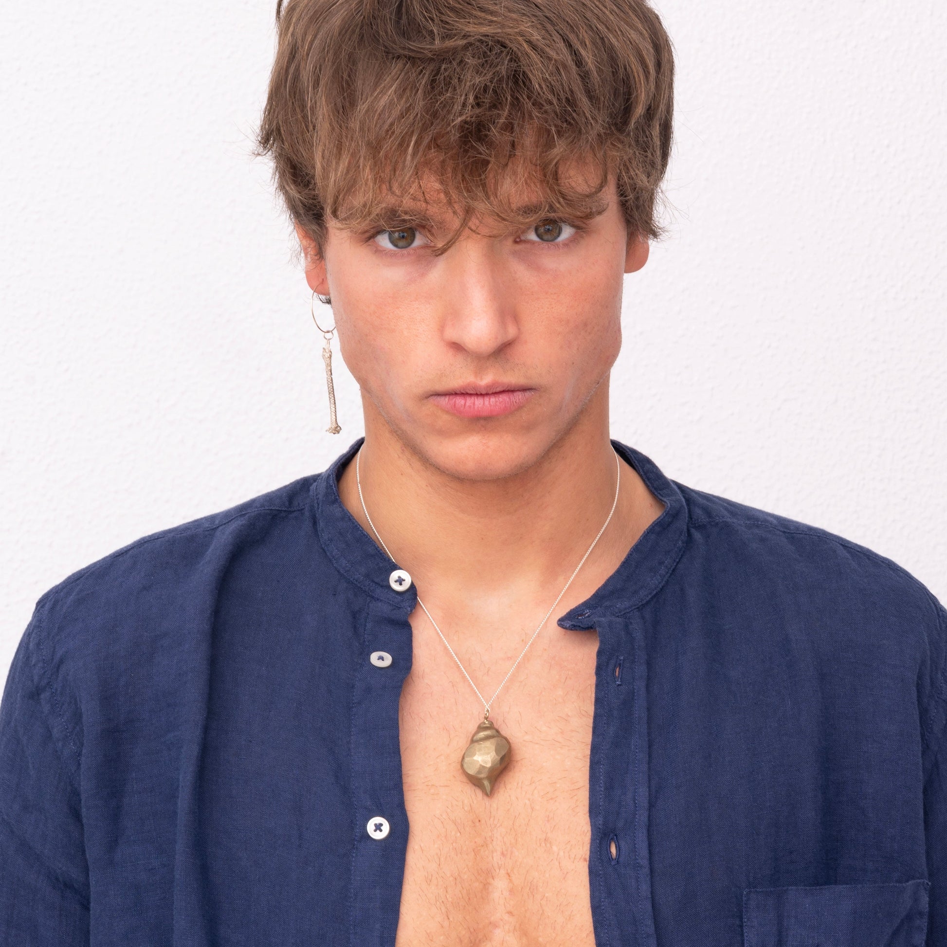 A person with short hair and a serious expression wears a blue unbuttoned shirt, an artisan-made necklace with a pendant, and a Thin Hoop Nautical Earring with Sterling Silver Rope, standing against a plain white background.