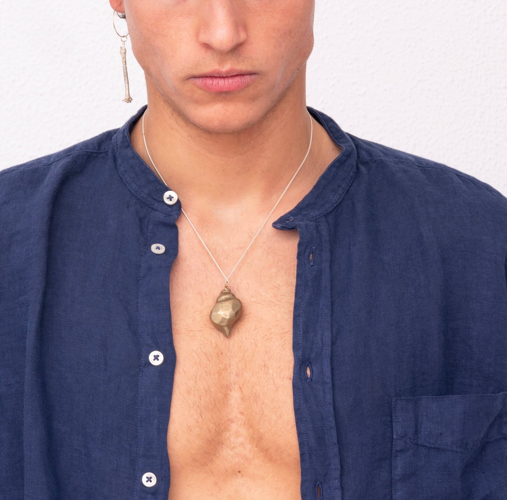 Young man with light brown hair wearing a navy blue shirt, a single earring, and the Nautical Handcrafted Seashell One of A Kind Pendant Nº 1 by Angela Lago poses against a plain white background.